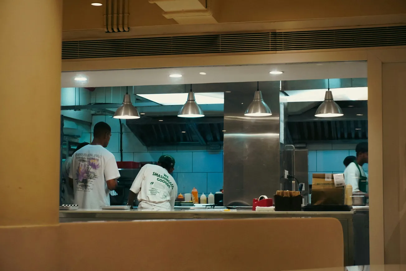 Restaurant kitchen with chefs in white shirts preparing food. Stainless steel surfaces, utensils, and condiment bottles are visible under bright overhead lights.