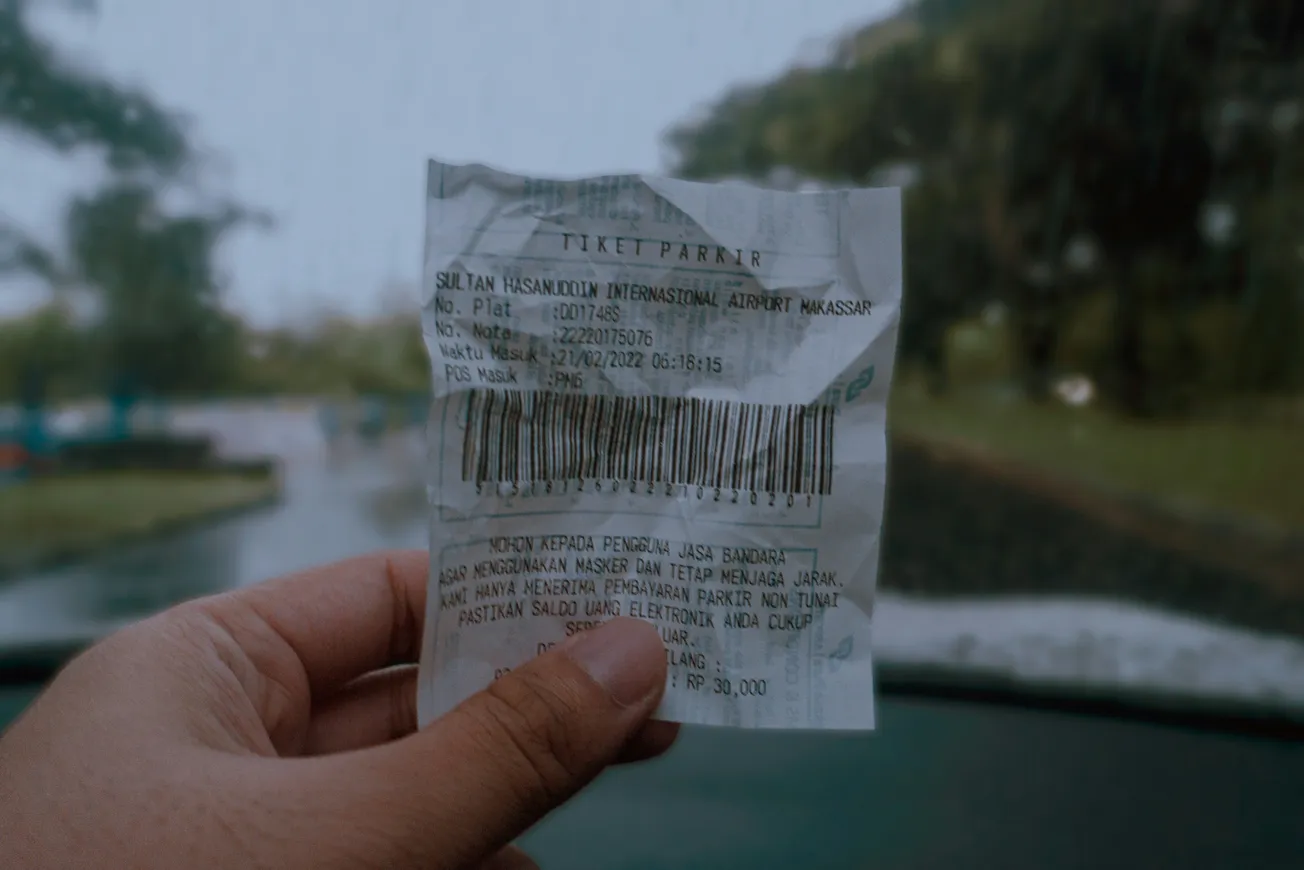 A hand holds a parking ticket for Sultan Hasanuddin International Airport. Blurry, rainy background of a road seen through a windshield. Moody atmosphere.