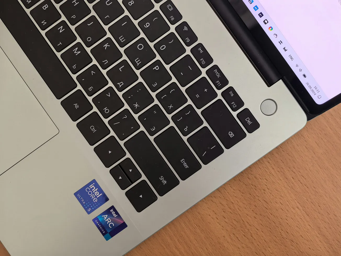 Close-up of a laptop keyboard with Cyrillic and English keys on a wooden desk. Intel Core and Arc stickers are visible near the touchpad.