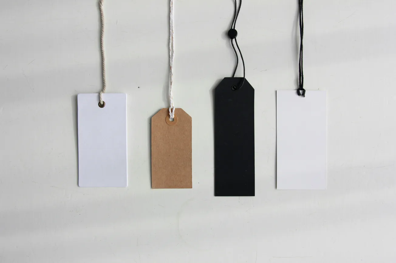 Four blank clothing hang tags in white, brown kraft, and black styles hanging by strings against a plain white background.