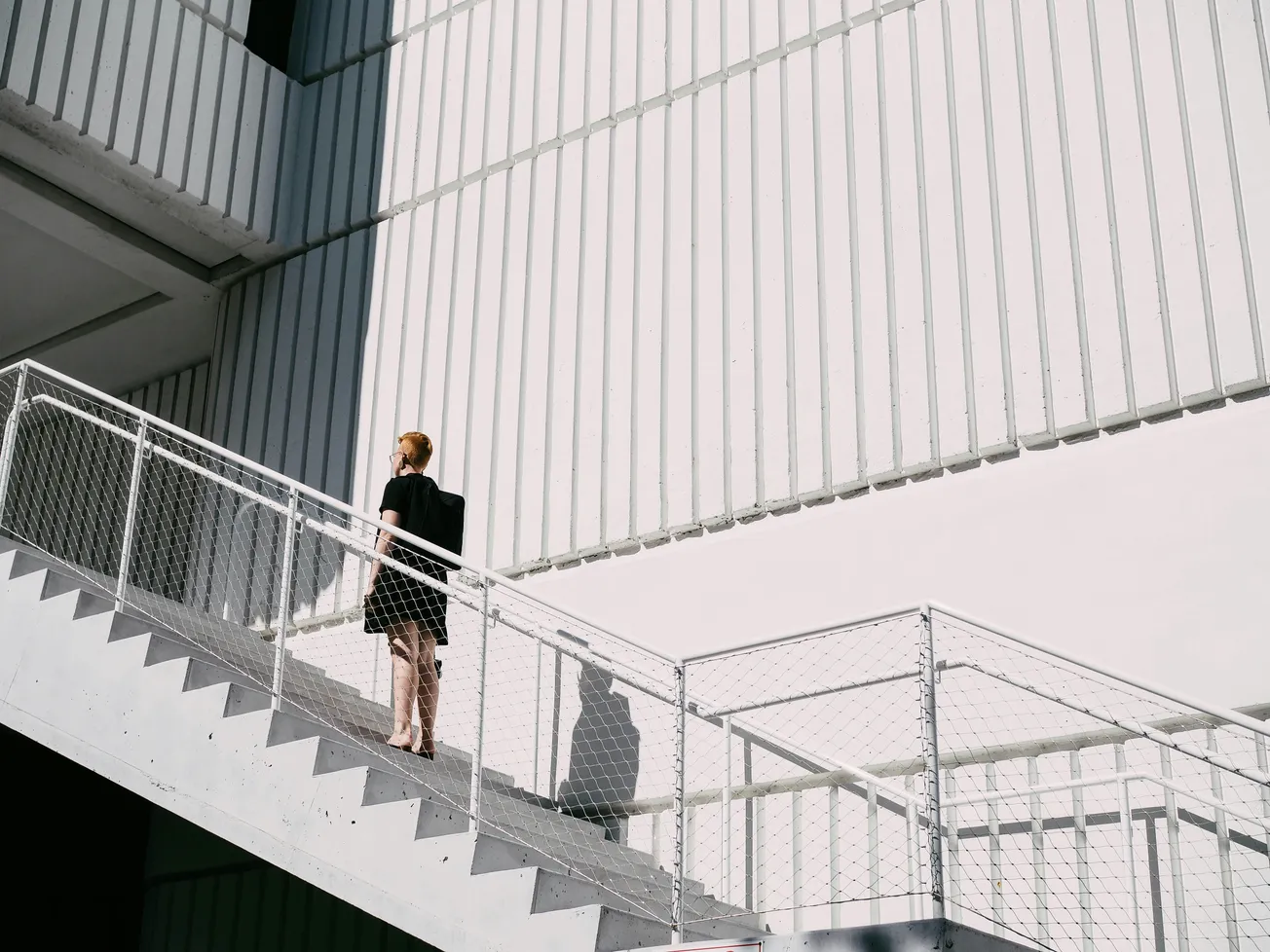 A person in a black dress ascends a sunlit, external staircase against a white, textured building. The scene feels minimalistic and modern.