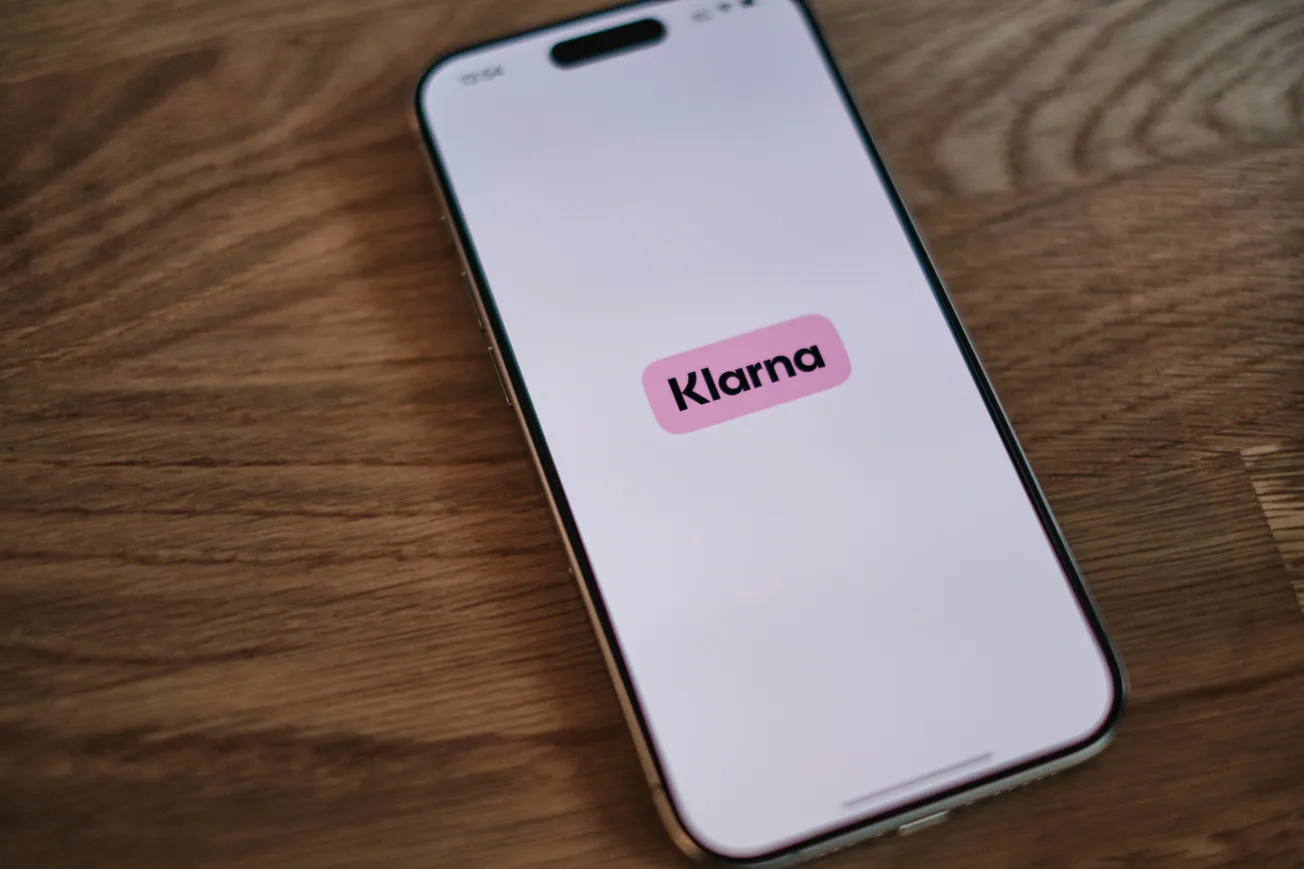 Smartphone displaying the Klarna app logo on its screen, resting on a wooden surface. The logo is pink, conveying a modern and sleek tone.