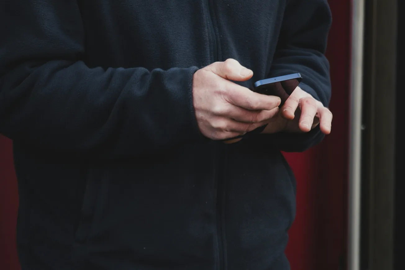 A person in a dark jacket uses a smartphone with both hands. The background is blurred, creating a focused and contemplative mood.