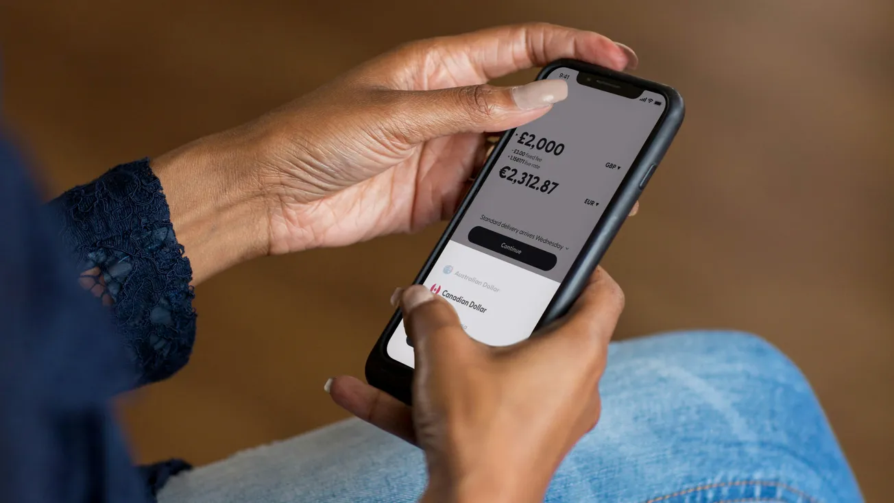 A person holds a smartphone, displaying a banking app with currency exchange details. The screen shows options to donate. The scene feels modern and focused.
