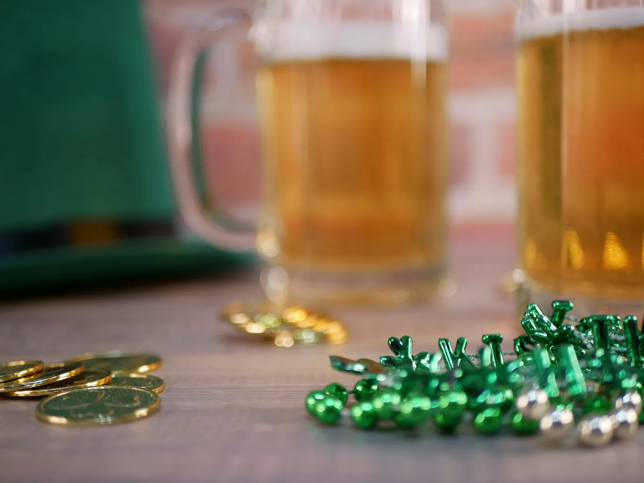 St. Patrick's Day theme with green beads, gold coins, frothy beer mugs, and a green hat. Festive and celebratory atmosphere against a blurred background.