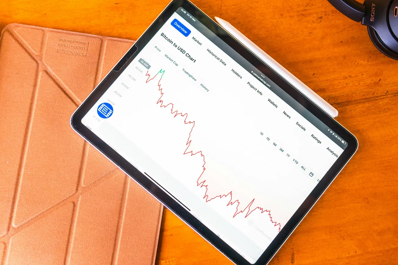 A tablet displays a Bitcoin to USD price chart showing a significant downward trend on a white background, resting on a wooden desk next to a stylus and headphones.