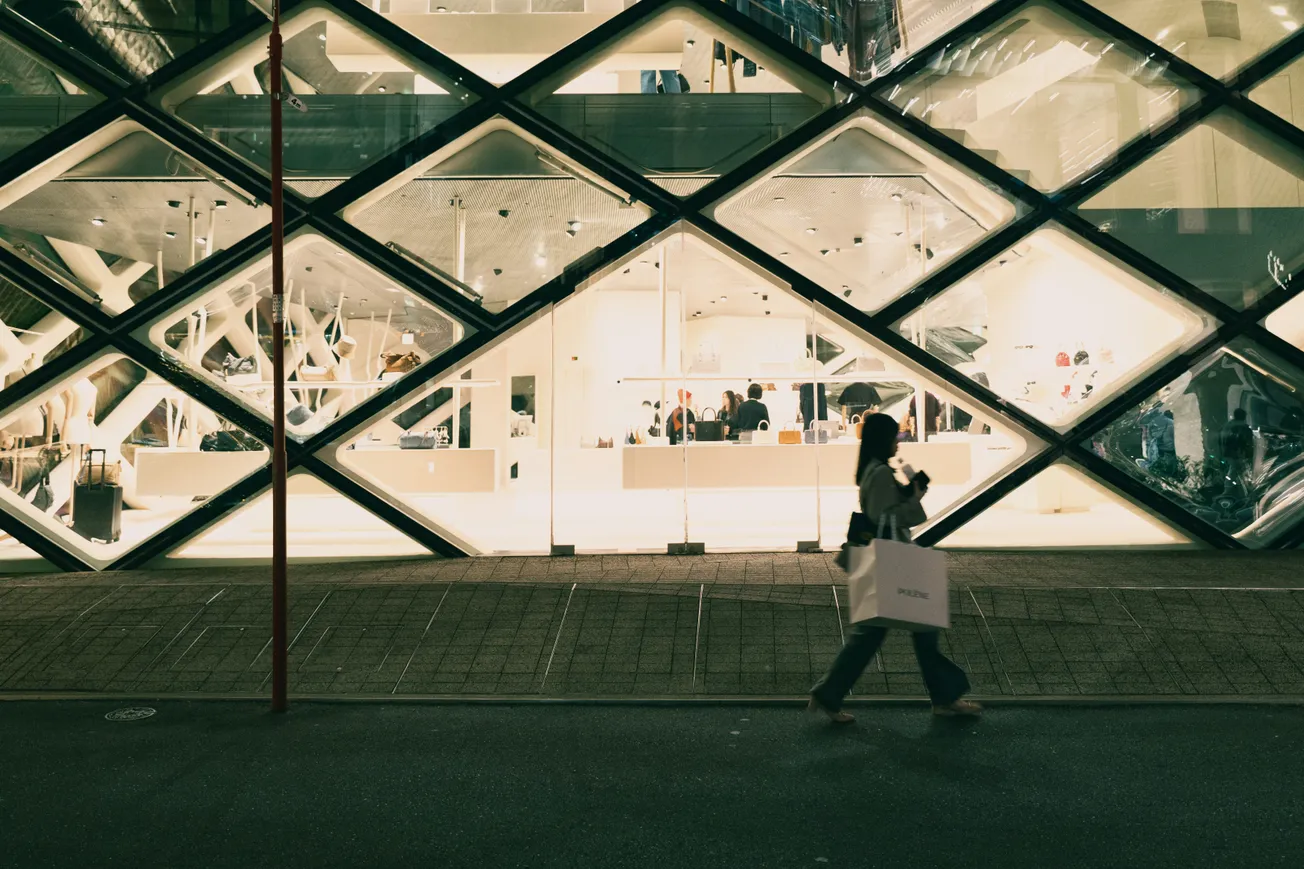 A woman walks past a modern building with diamond-patterned glass. Inside, a bright, minimalistic store is visible. The scene is calm and urban.