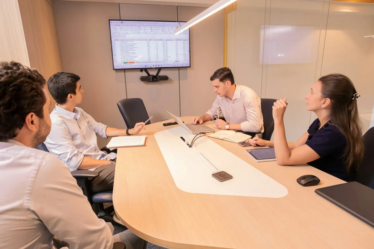 Four people in a conference room discuss data displayed on a wall-mounted screen. The setting is professional, focused, and collaborative.