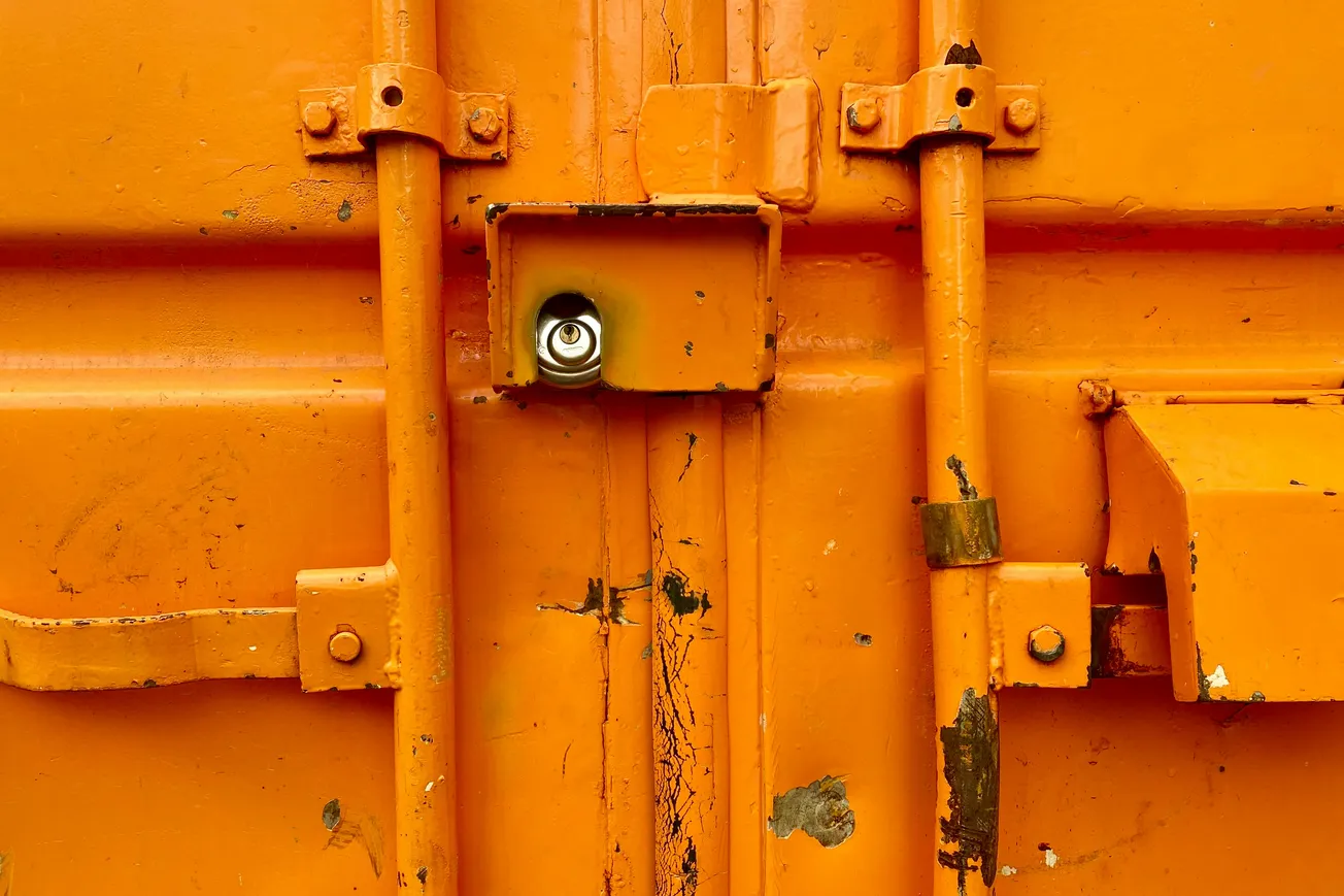 Close-up of an orange metal door with visible rust and scratches, featuring vertical and horizontal bars and a shiny silver lock at the center.