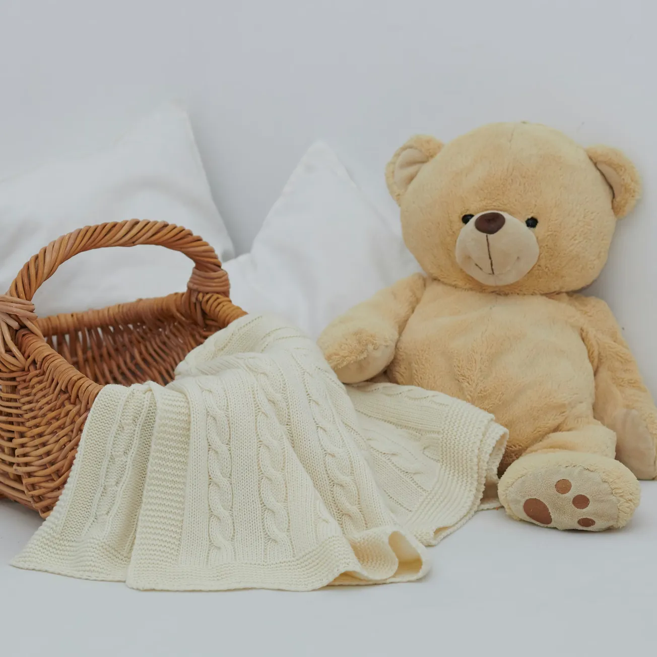 A plush teddy bear sits beside a wicker basket draped with a cream-colored cable knit blanket. The scene is cozy and inviting, set against a white background.