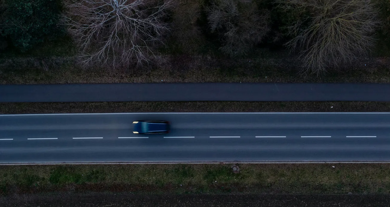 Aerial view of a lone black car driving on a two-lane road flanked by bare trees and grass, conveying speed and isolation.
