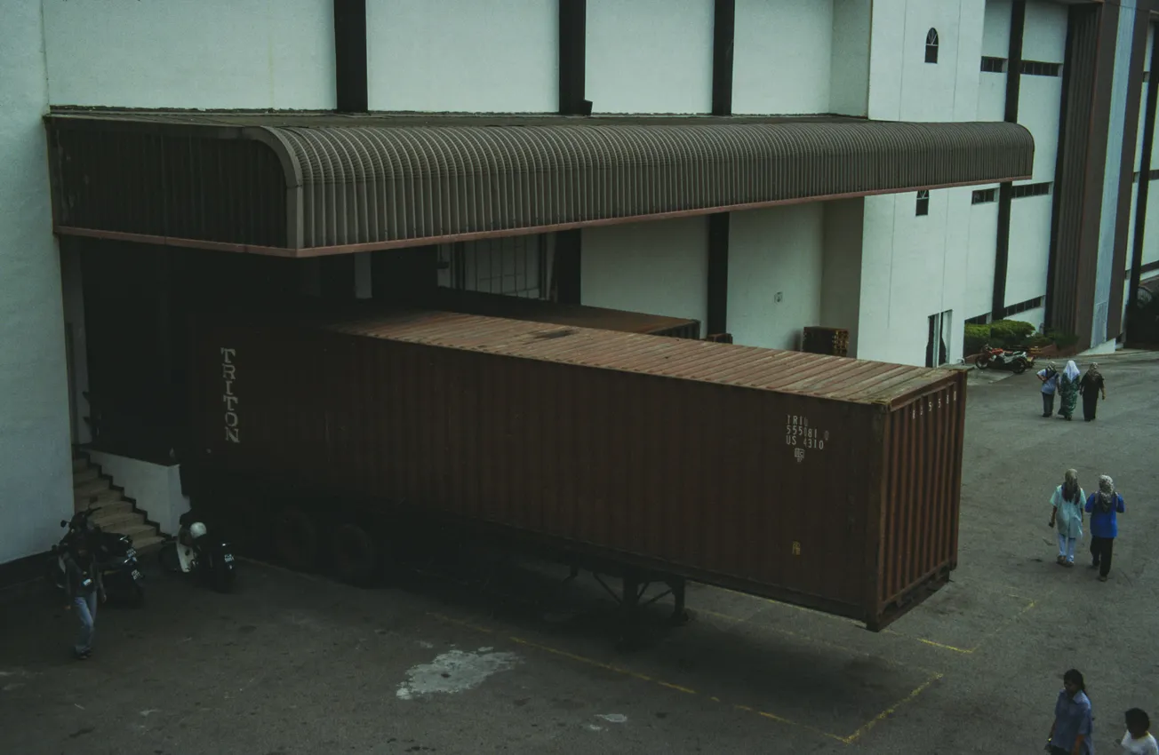 A large shipping container truck is docked at a warehouse loading bay under a corrugated metal roof. Four people are walking nearby. The mood is industrial and busy.