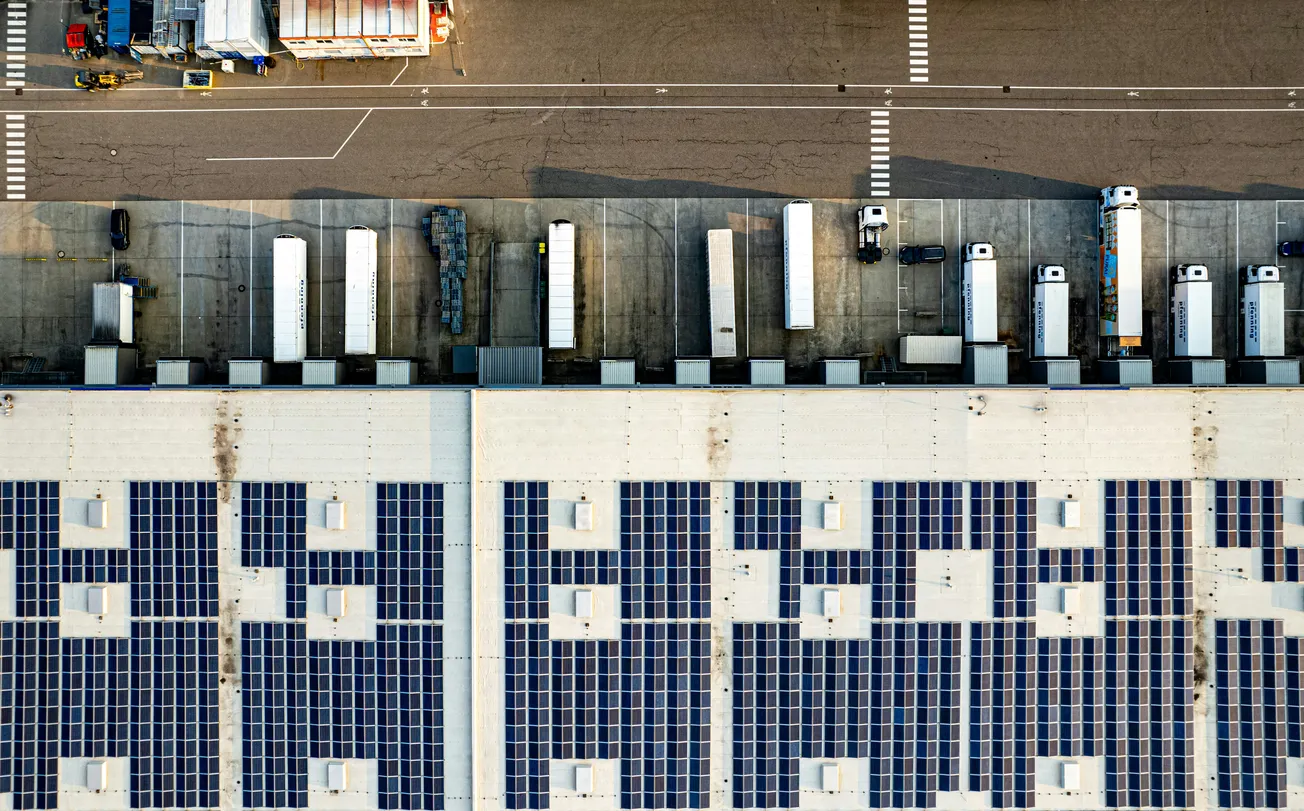 Aerial view of a large building with grid-patterned solar panels on the roof. Adjacent are parked semi-trucks. The scene conveys efficiency and sustainability.