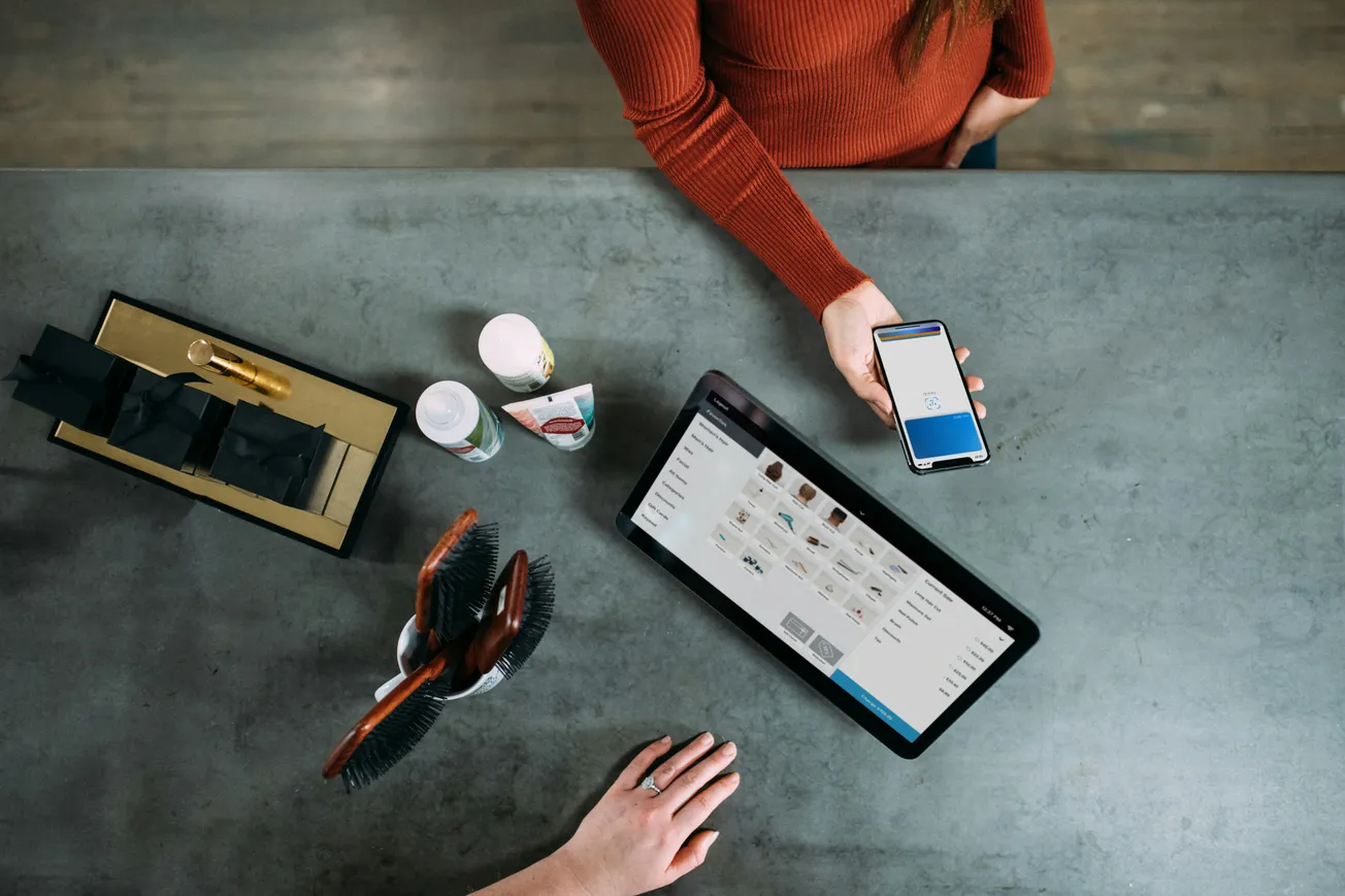 Overhead view of two people at a table, one holding a phone with a payment app, the other by a tablet displaying products.