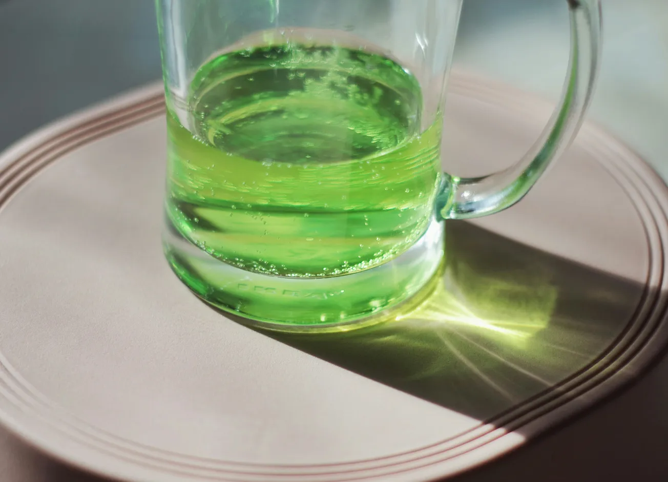 A glass mug filled with bright green liquid sits on a light beige circular surface. Sunlight creates a vivid reflective pattern beside the mug.