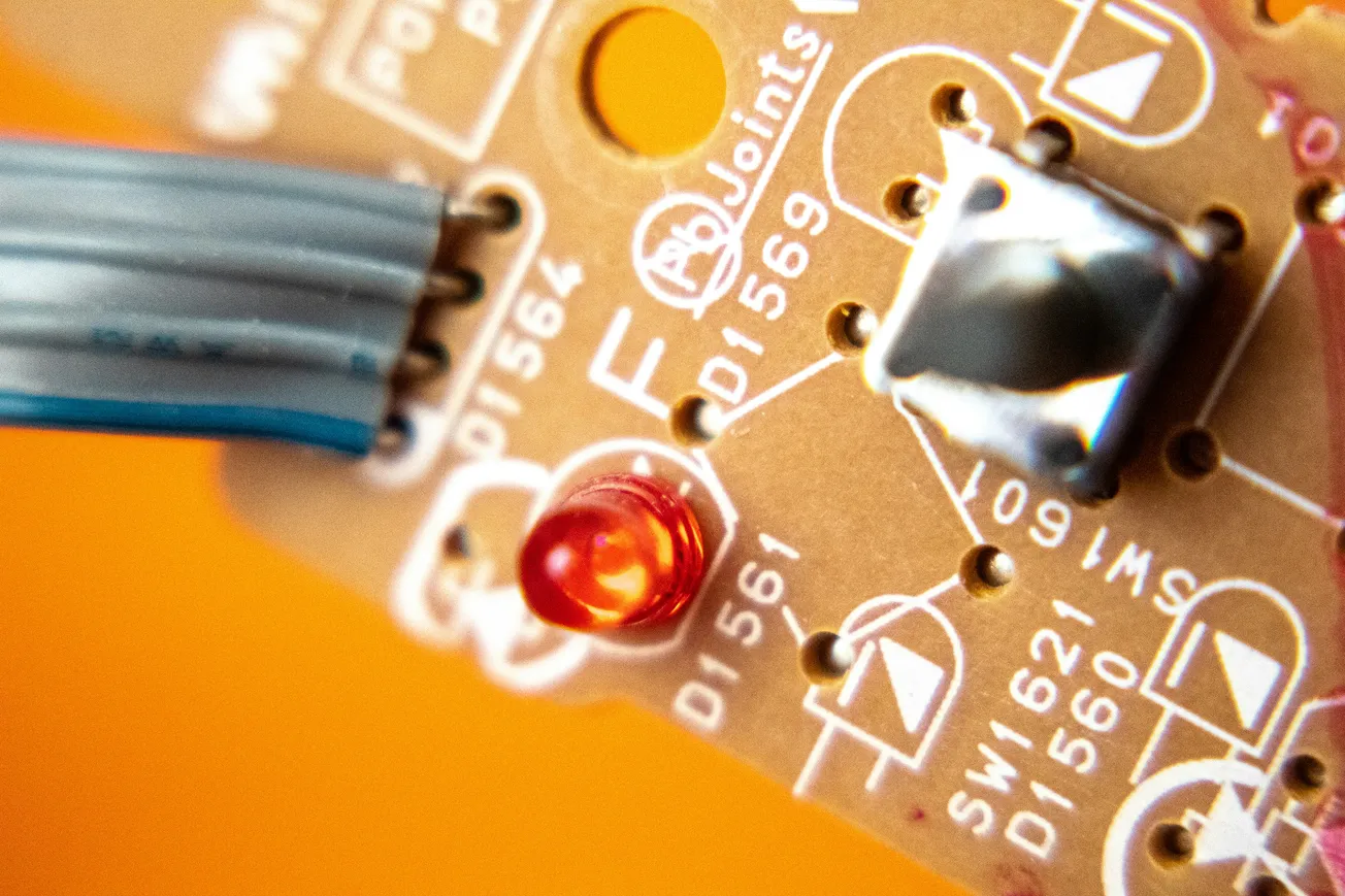 Close-up of a circuit board with a glowing red LED, blue cable, and metallic components on a vibrant orange background, conveying a tech-focused tone.