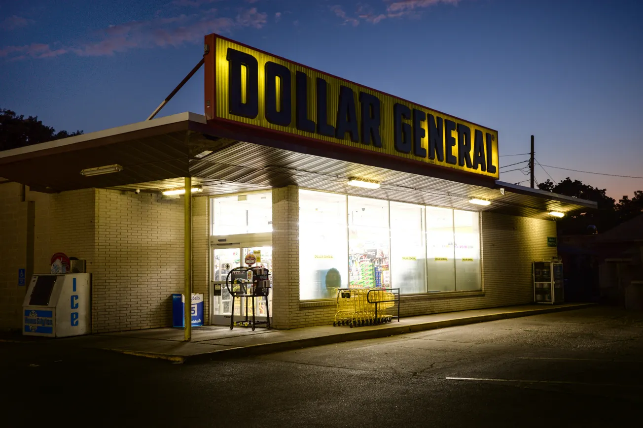 A brightly lit Dollar General store at dusk. The neon sign glows against the evening sky, casting warm light over yellow shopping carts outside.