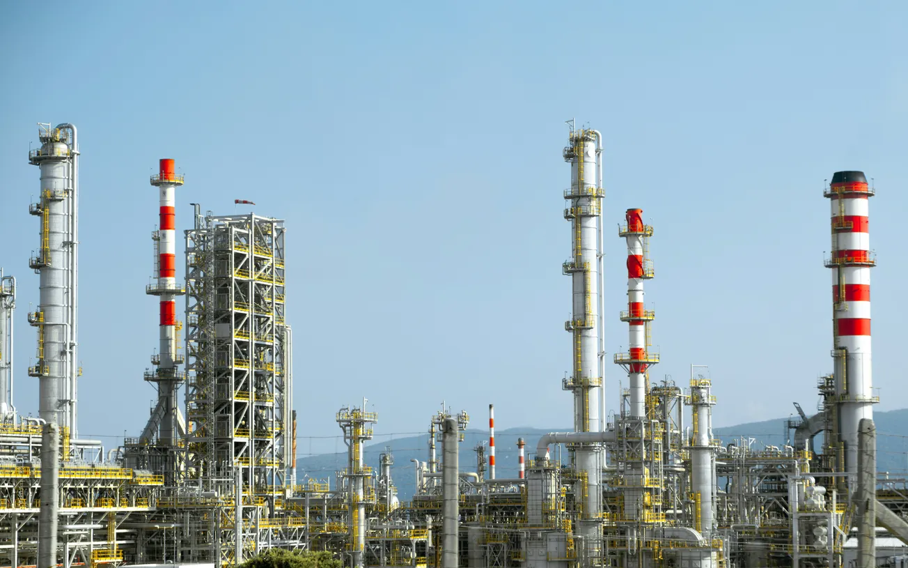Industrial refinery with tall chimneys featuring red and white stripes, set against a clear blue sky. Metal structures complexly intertwine.