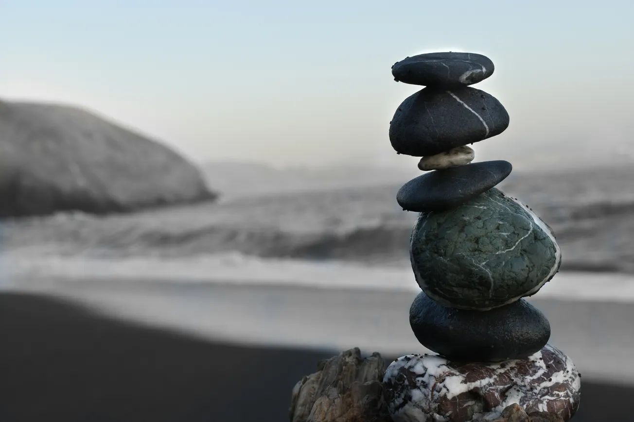 Stacked stones balanced on driftwood on a serene beach, with blurred waves and rocky coast in the background, creating a peaceful, harmonious scene.