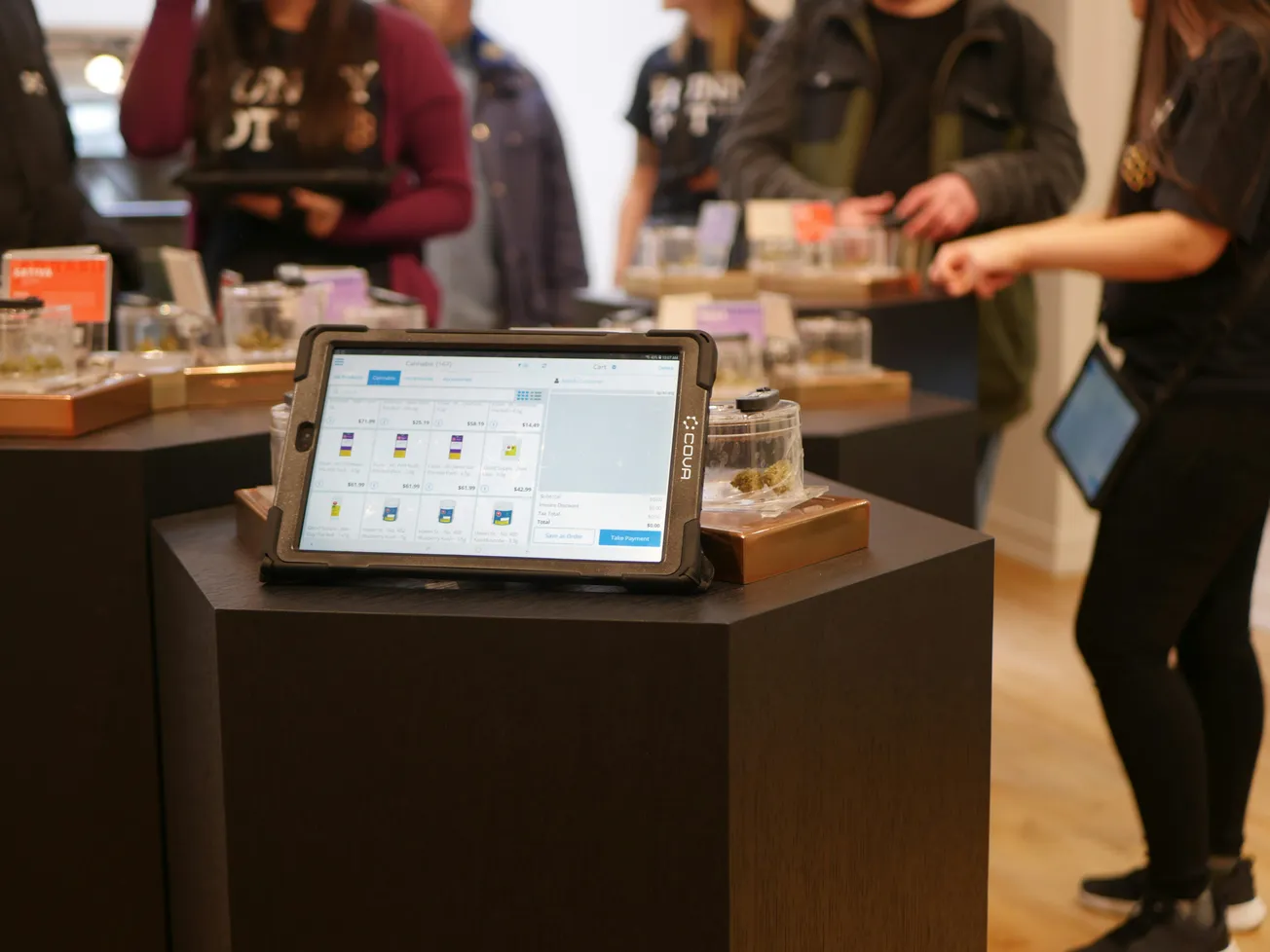 Tablet displaying product information on a black pedestal, surrounded by people in casual clothes, suggests a modern store setting.