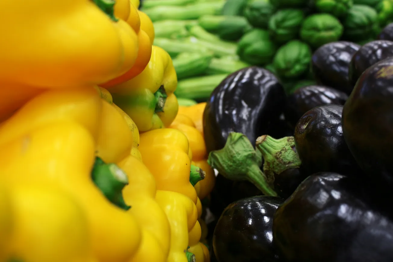 A vibrant market display with stacks of yellow bell peppers, glossy eggplants, and green leafy vegetables, conveying freshness and abundance.