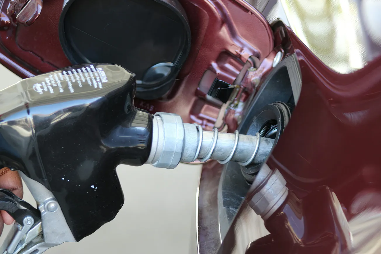 A close-up of a black gas nozzle inserted into a car's fuel tank. The car is maroon, and the shiny nozzle reflects light, conveying utility and focus.