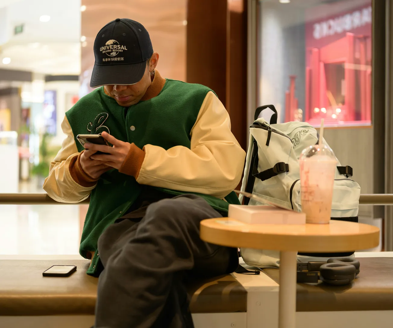 Man in a green jacket and cap sits in a mall, focused on his phone. A white backpack and drink are nearby, creating a casual, relaxed mood.