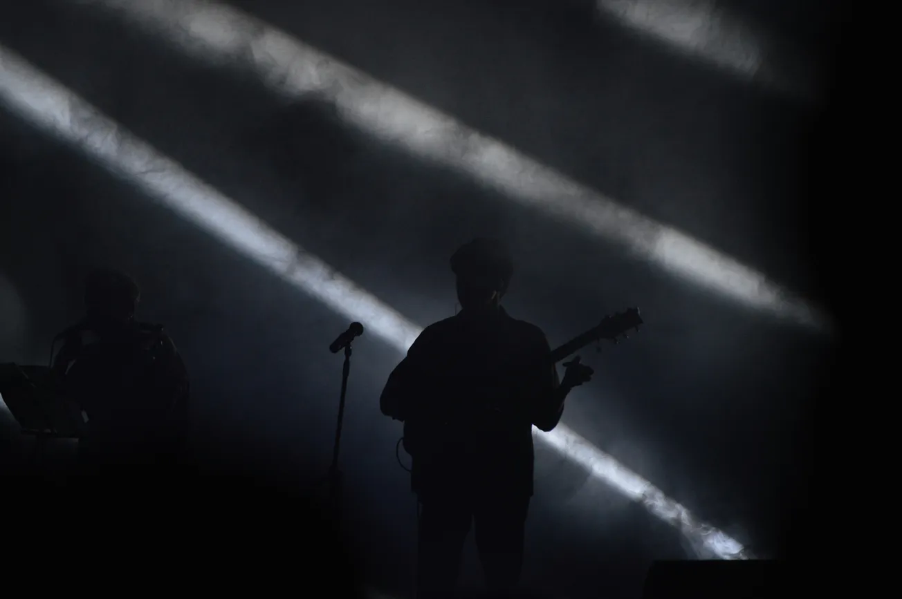 Silhouetted musician playing guitar on a dark stage with dramatic, diagonal beams of light cutting through fog. Moody and atmospheric concert scene.