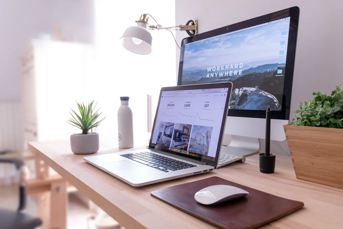 A modern workspace with a wooden desk featuring an open laptop displaying graphs, an external monitor with "Work Hard Anywhere," a lamp, plant, and mouse.