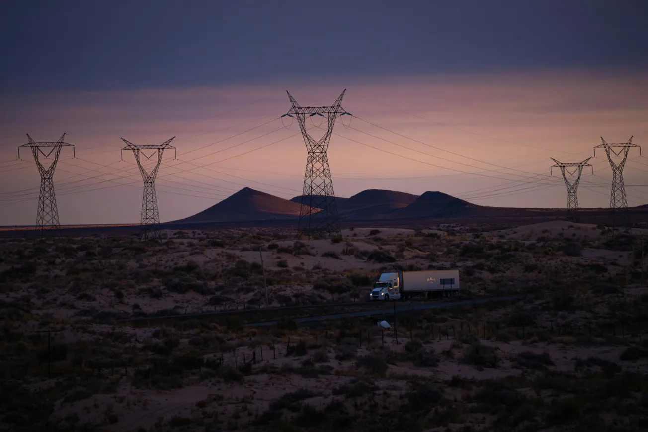 A lone truck drives through a desert landscape at dusk, flanked by towering power lines. The sky fades from purple to pink over distant hills, creating a serene atmosphere.