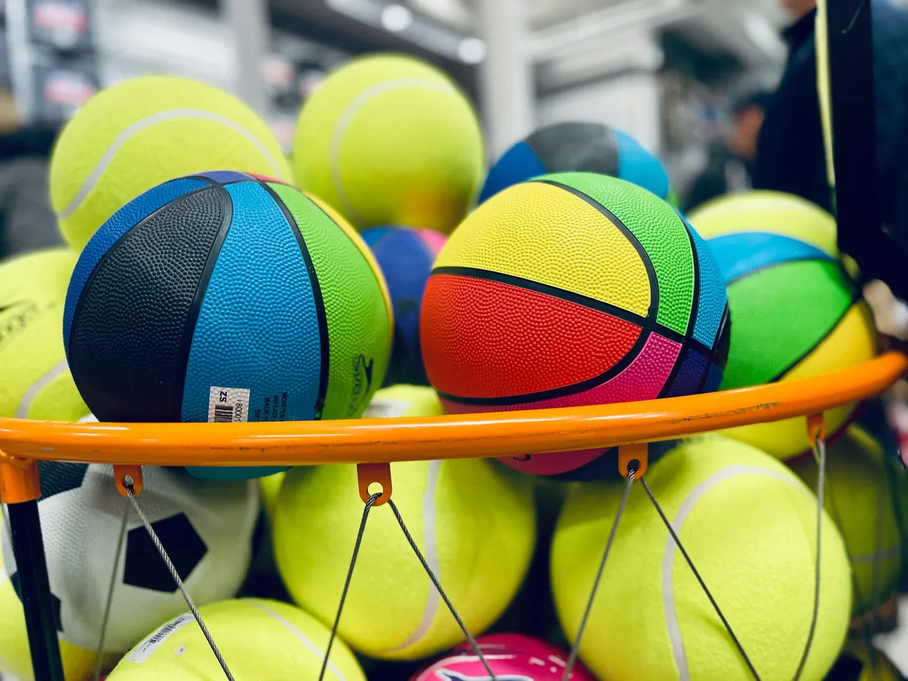 A pile of colorful sports balls in a store display, including neon basketballs and yellow tennis balls, with a vibrant and energetic atmosphere.
