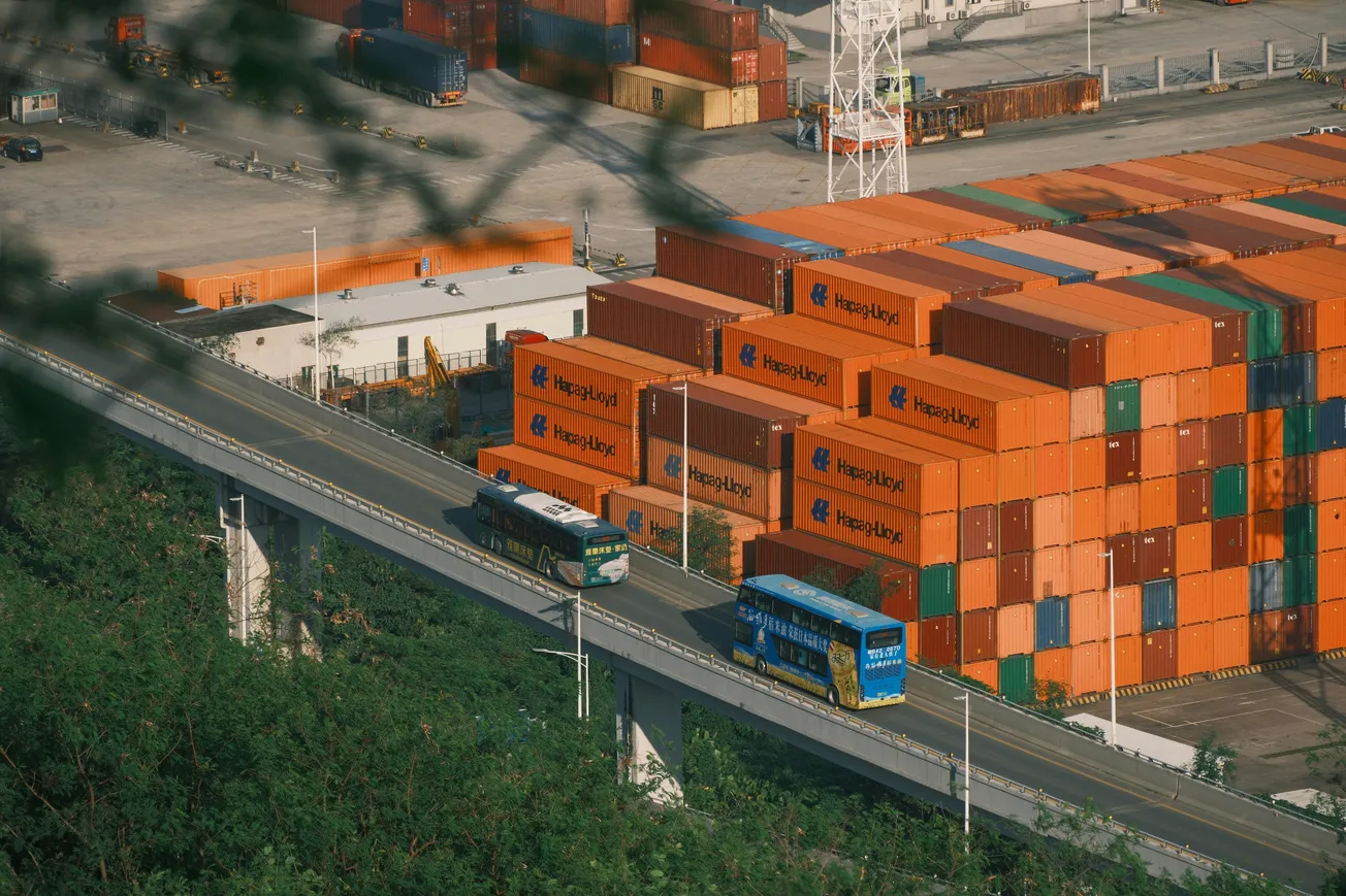 Aerial view of a shipping yard with stacked orange containers marked "Hapag-Lloyd." Two buses travel on an elevated road amidst lush greenery.