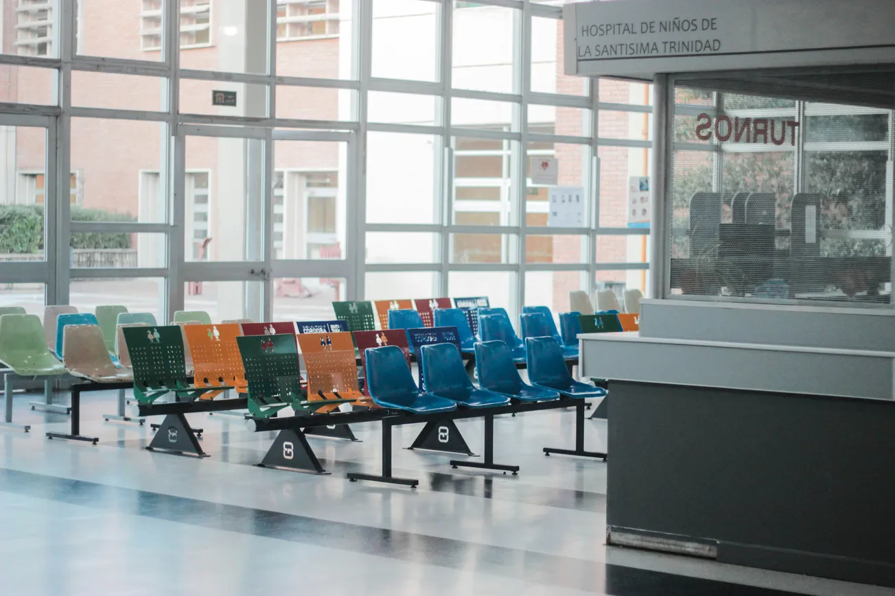 Bright hospital waiting area with large windows, featuring rows of colorful empty chairs. Natural light fills the space, creating a calm atmosphere.