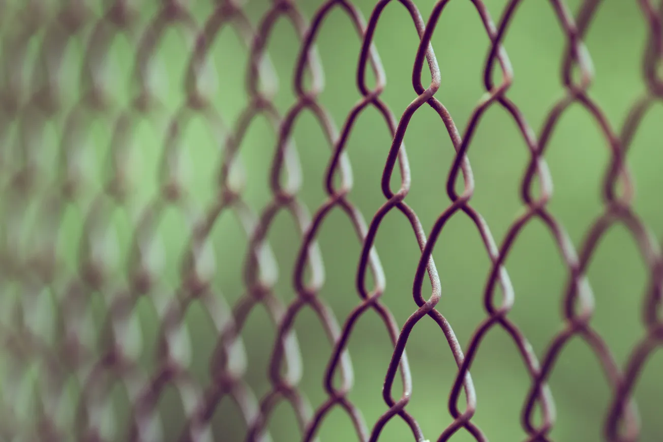 Close-up of a rusty chain-link fence, with diamond shapes, against a blurred green background, conveying a sense of isolation and simplicity.