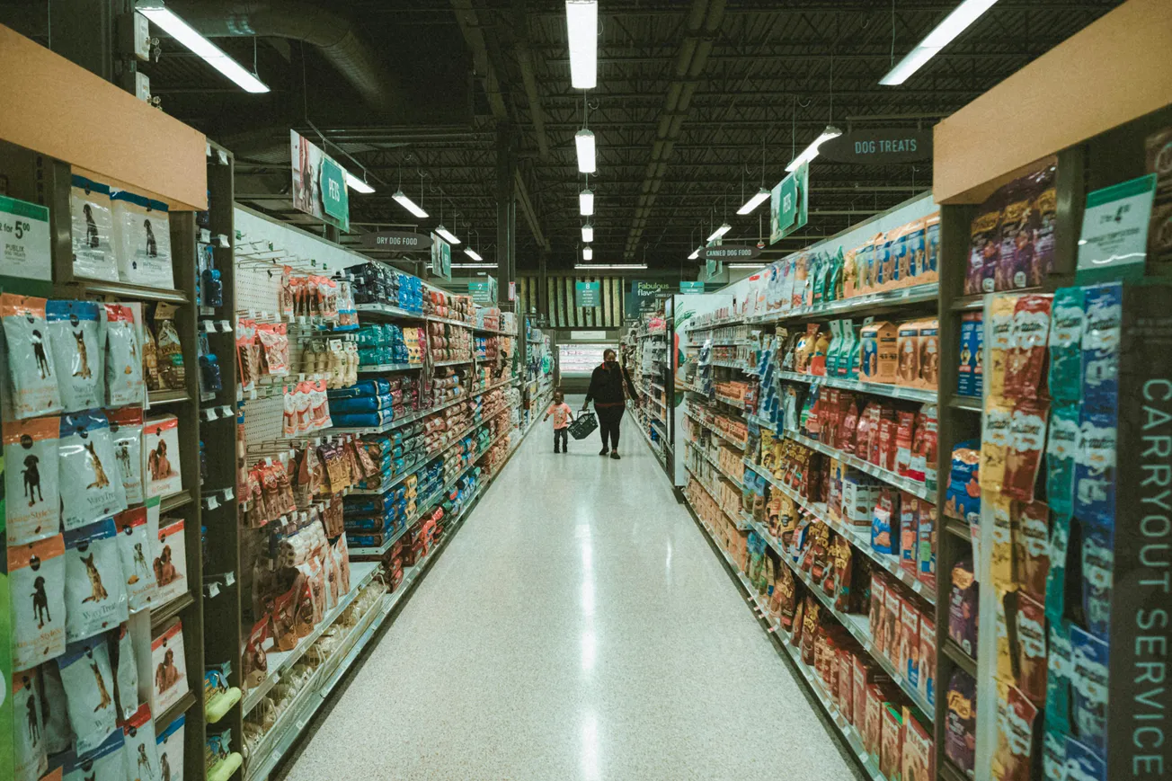 Wide supermarket aisle with pet food on both sides. A person and child walk down the brightly lit aisle, creating a calm, everyday shopping scene.