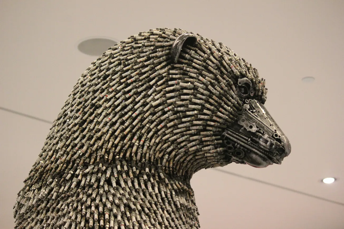 Sculpture of a bear made from metallic elements, resembling bolts, displayed indoors. The bear's expression is solemn, with intricate detailing.