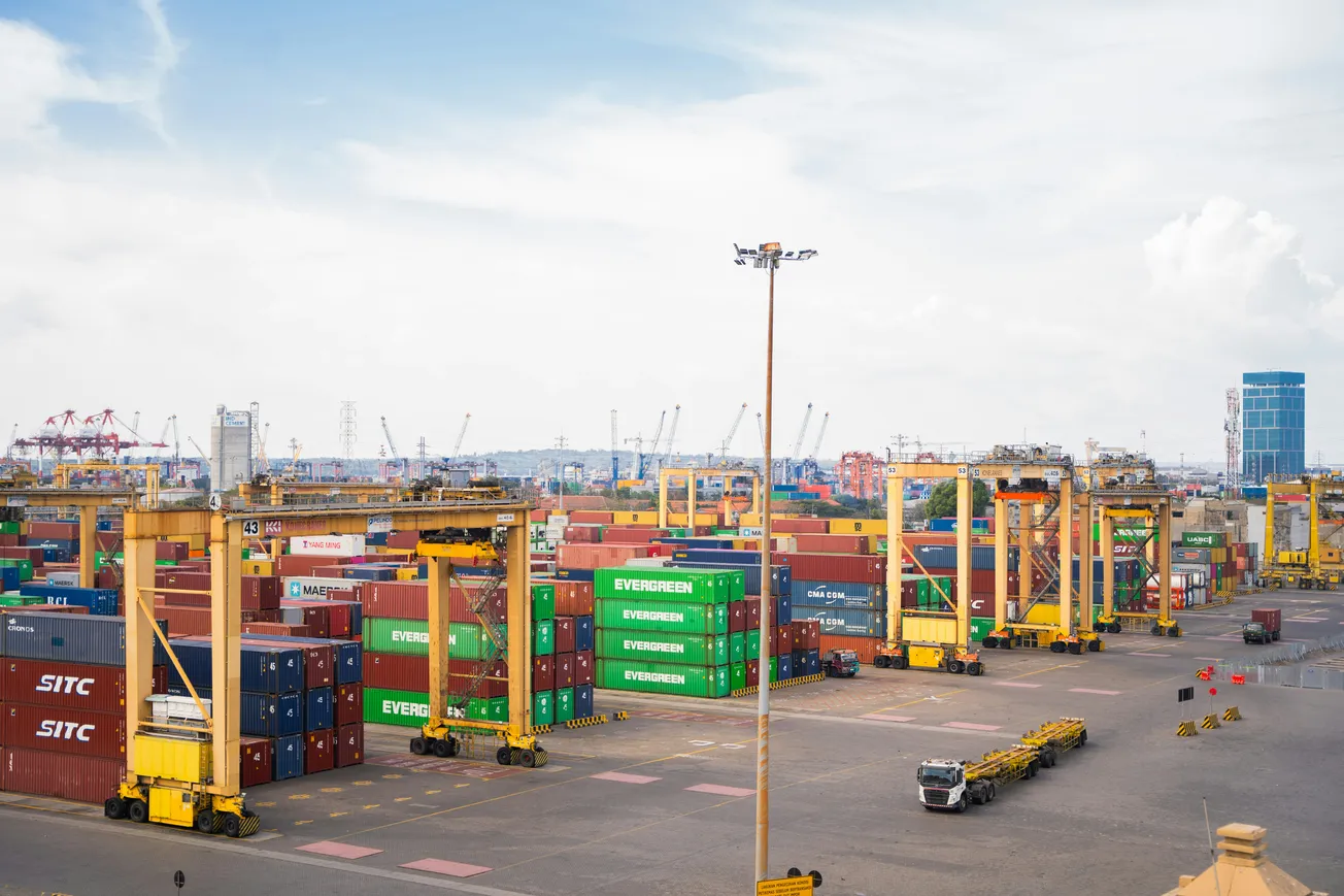 A bustling port filled with stacked colorful shipping containers and large yellow cranes. Trucks are visible, conveying a busy, industrious atmosphere.