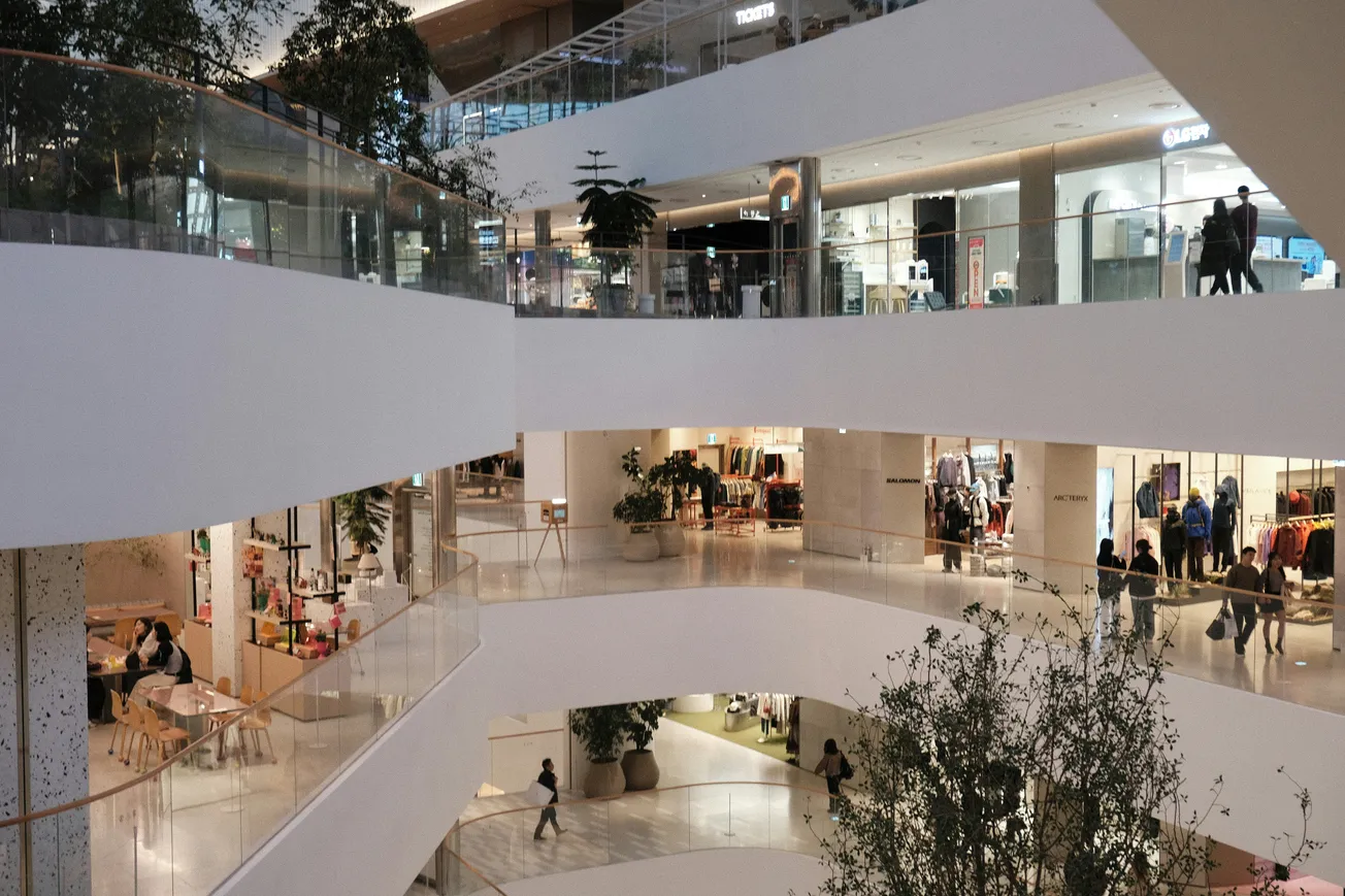 Modern multi-level shopping mall with sleek white railings, various store fronts, and shoppers. Indoor trees add greenery. Bright, bustling atmosphere.