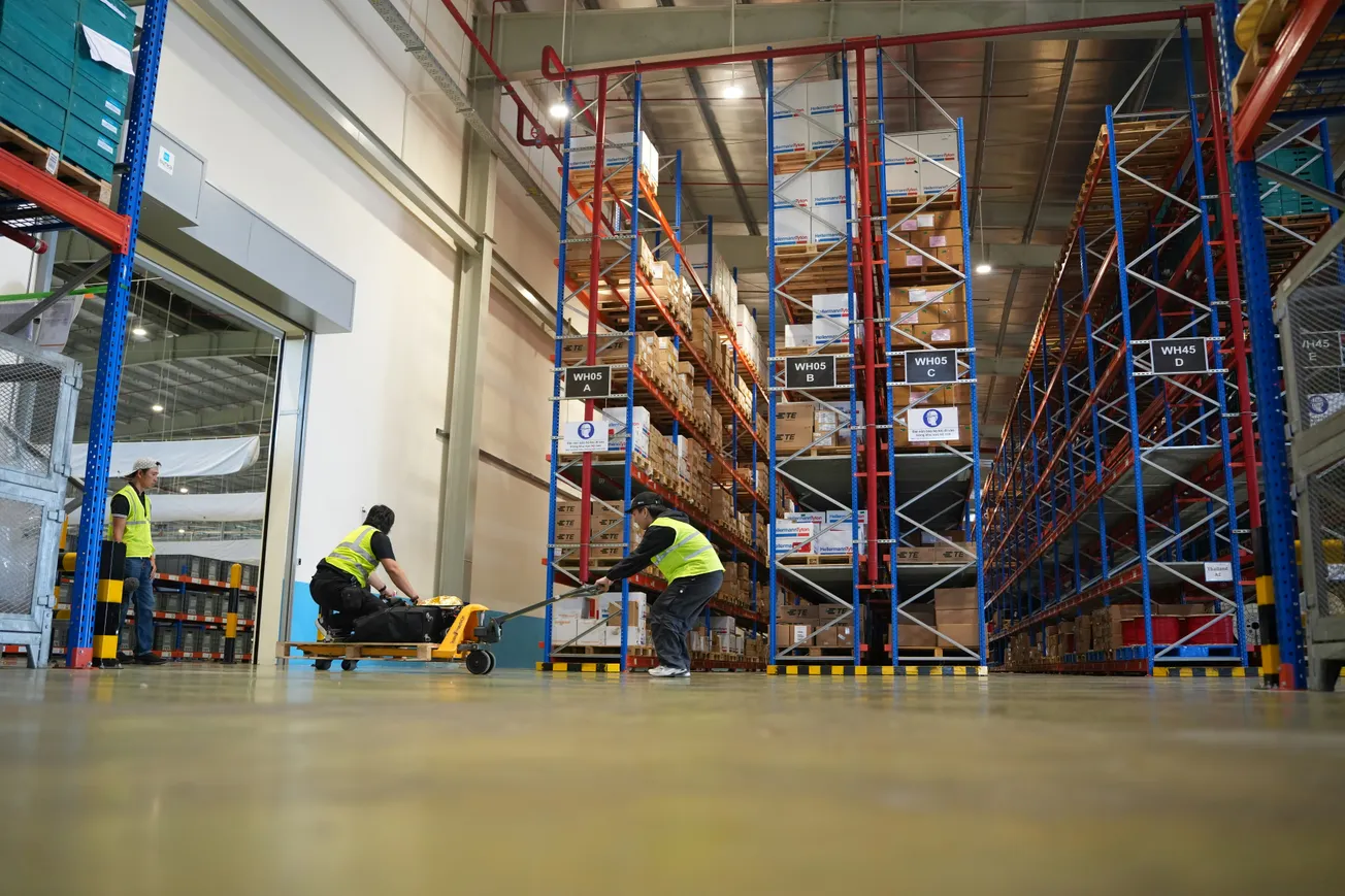 Warehouse scene with high racks of boxes. Two workers in yellow vests maneuver a hand pallet truck. A third worker watches, conveying teamwork.