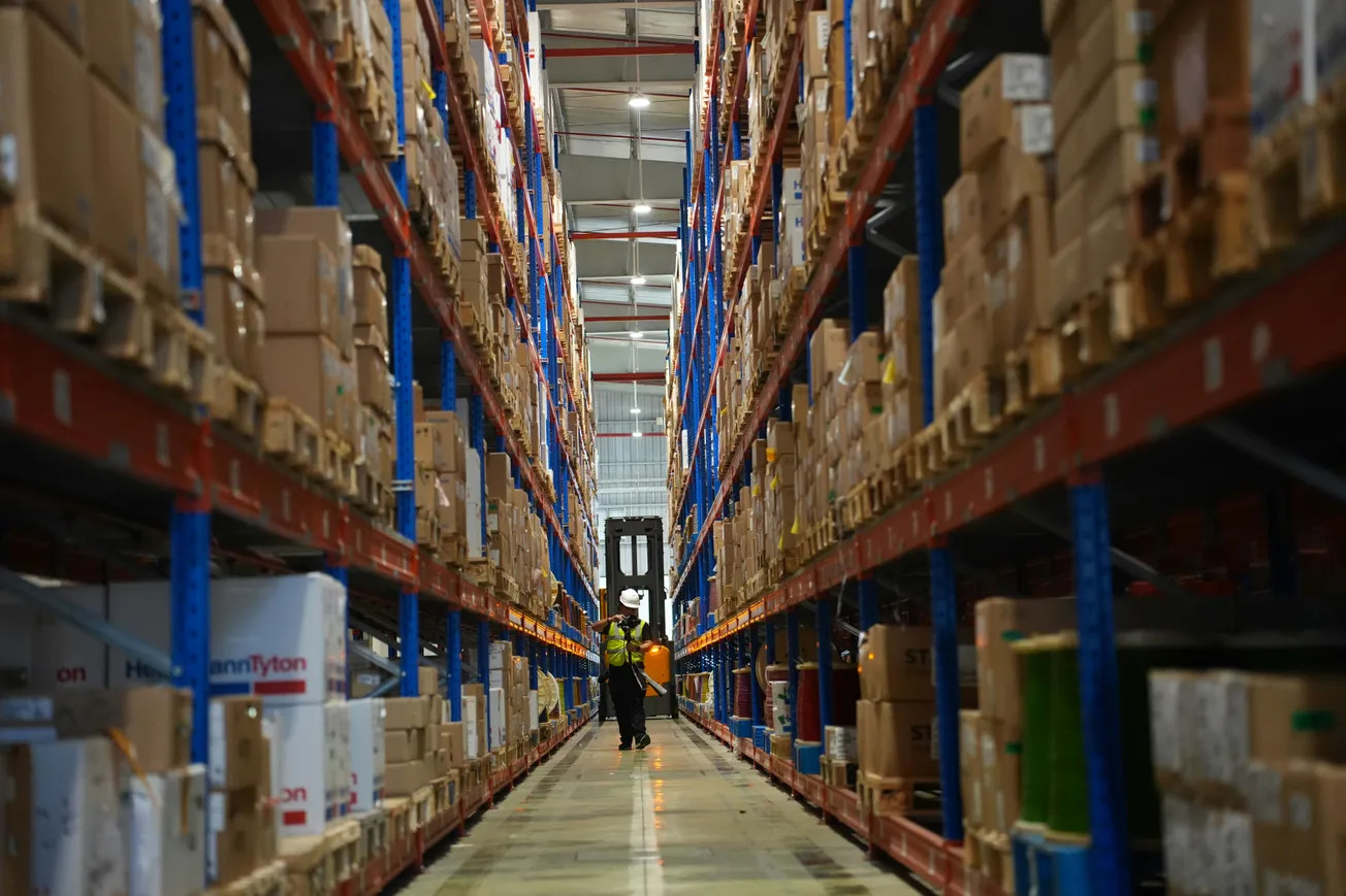 Wide-angle view of a large warehouse aisle filled with stacked boxes on high shelves. A person in a safety vest and helmet walks beside a forklift.