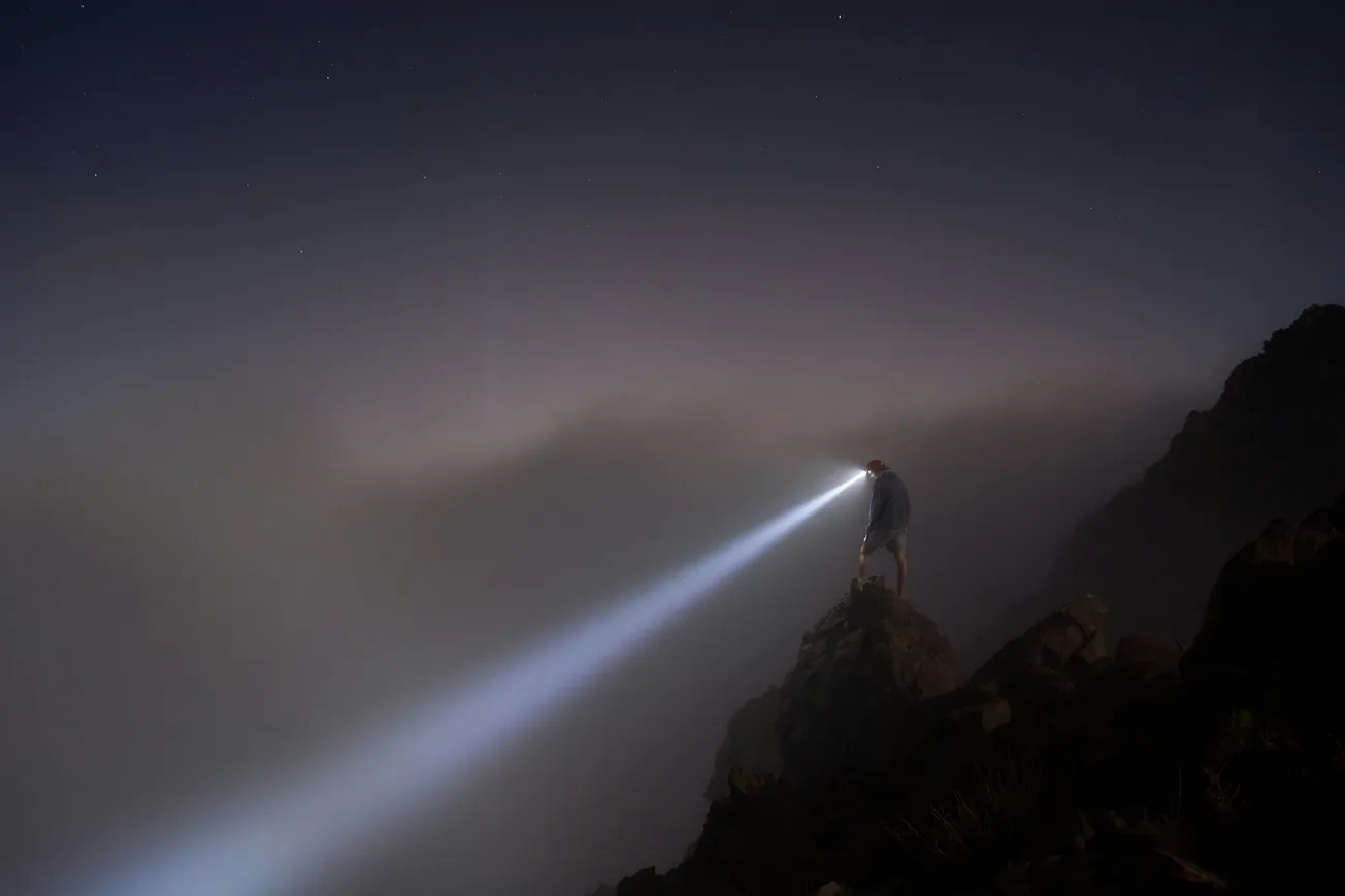 A person stands on a rocky cliff at night, shining a bright flashlight into foggy darkness.