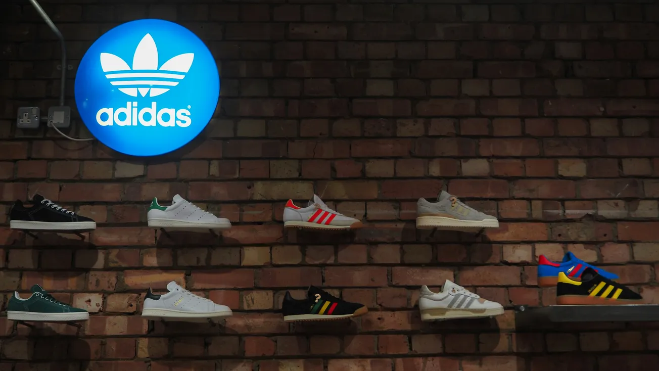 A brick wall displays various Adidas sneakers on shelves. Above, a glowing blue Adidas logo sign adds a modern touch to the vintage setting.
