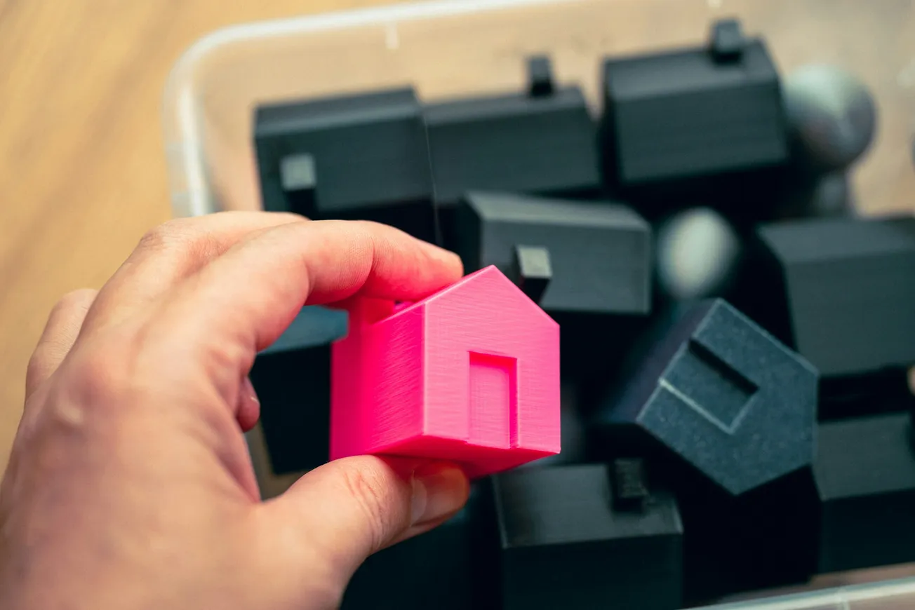 A hand holds a small, bright pink 3D-printed house model over a container filled with black house models, conveying contrast and focus.