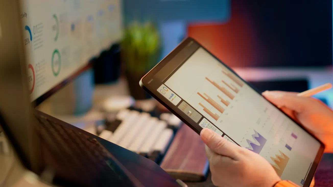 A person holds a tablet displaying bar and line graphs, with another screen showing charts in the background. The scene conveys a business and data analysis environment.