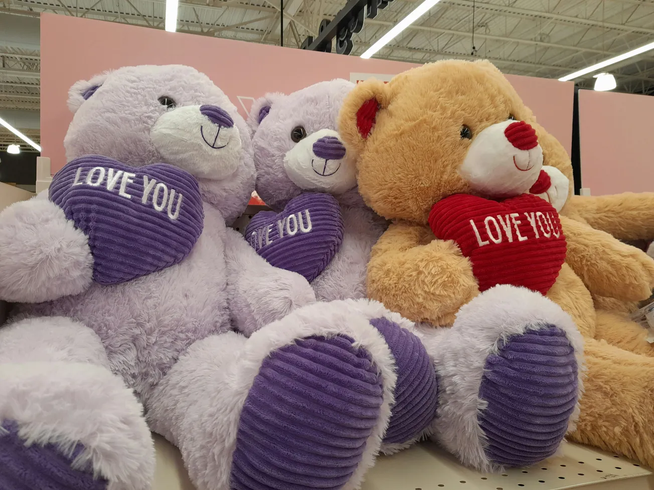Three plush teddy bears are on display in a store. Two are lavender holding purple "Love You" hearts, and one is tan holding a red heart.