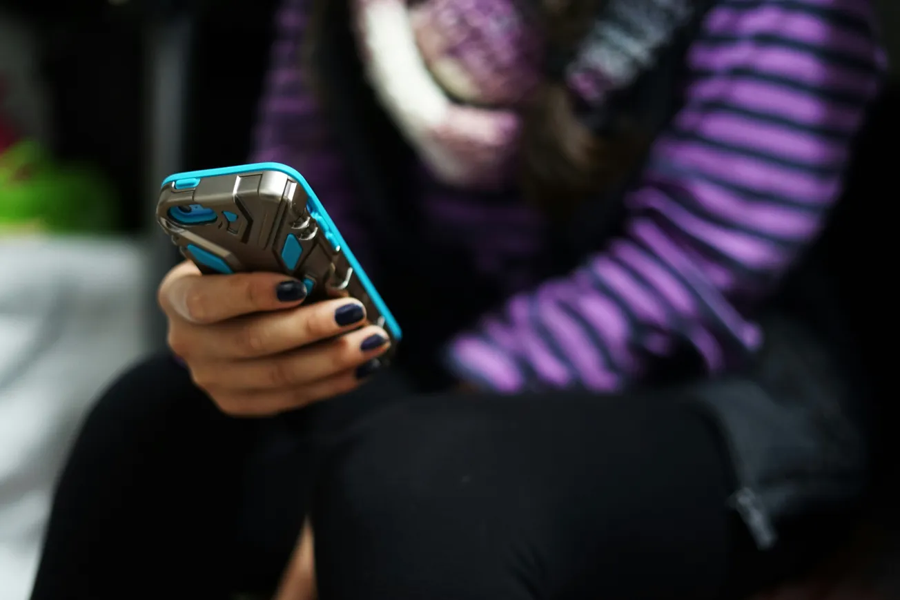 A person wearing a purple and black striped sweater and scarf is holding a smartphone with a rugged blue and gray case, focused on the screen.