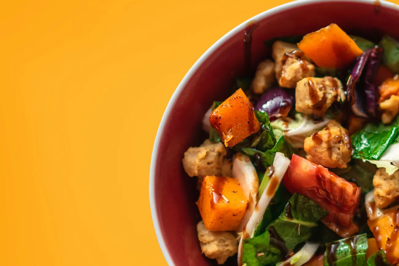 A vibrant salad in a red bowl against an orange background features roasted squash, leafy greens, tofu pieces, and red cabbage, creating a fresh and colorful dish.