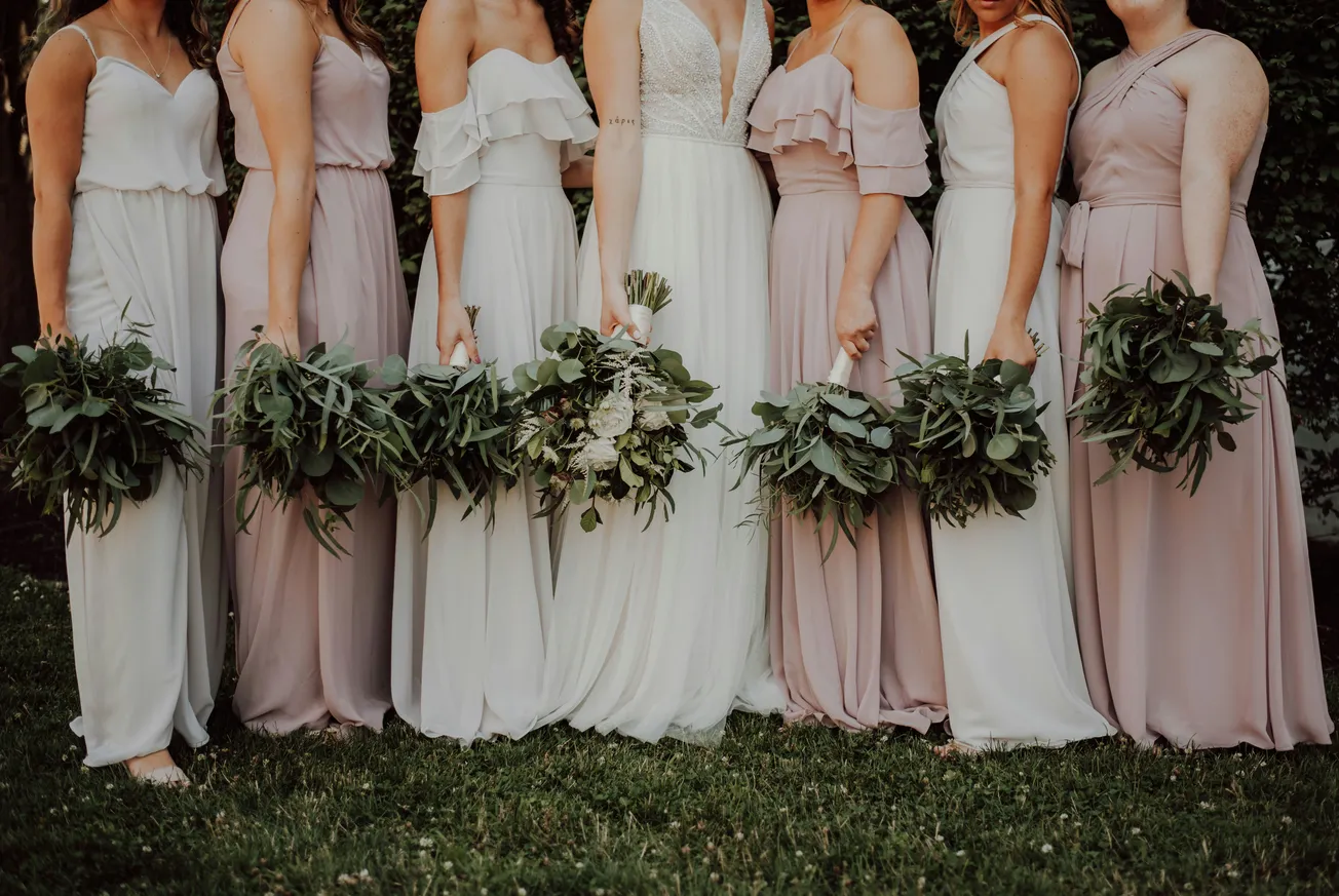 Bridal party in pastel dresses holding leafy bouquets, standing on grass. Elegance and harmony conveyed through soft colors and natural elements.