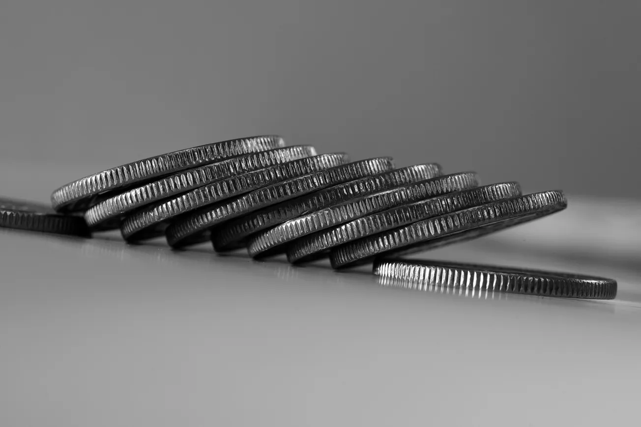 A stack of silver coins lies in a slightly tilted arrangement on a smooth surface, in black and white. The coins' edges are textured, creating a sense of balance.