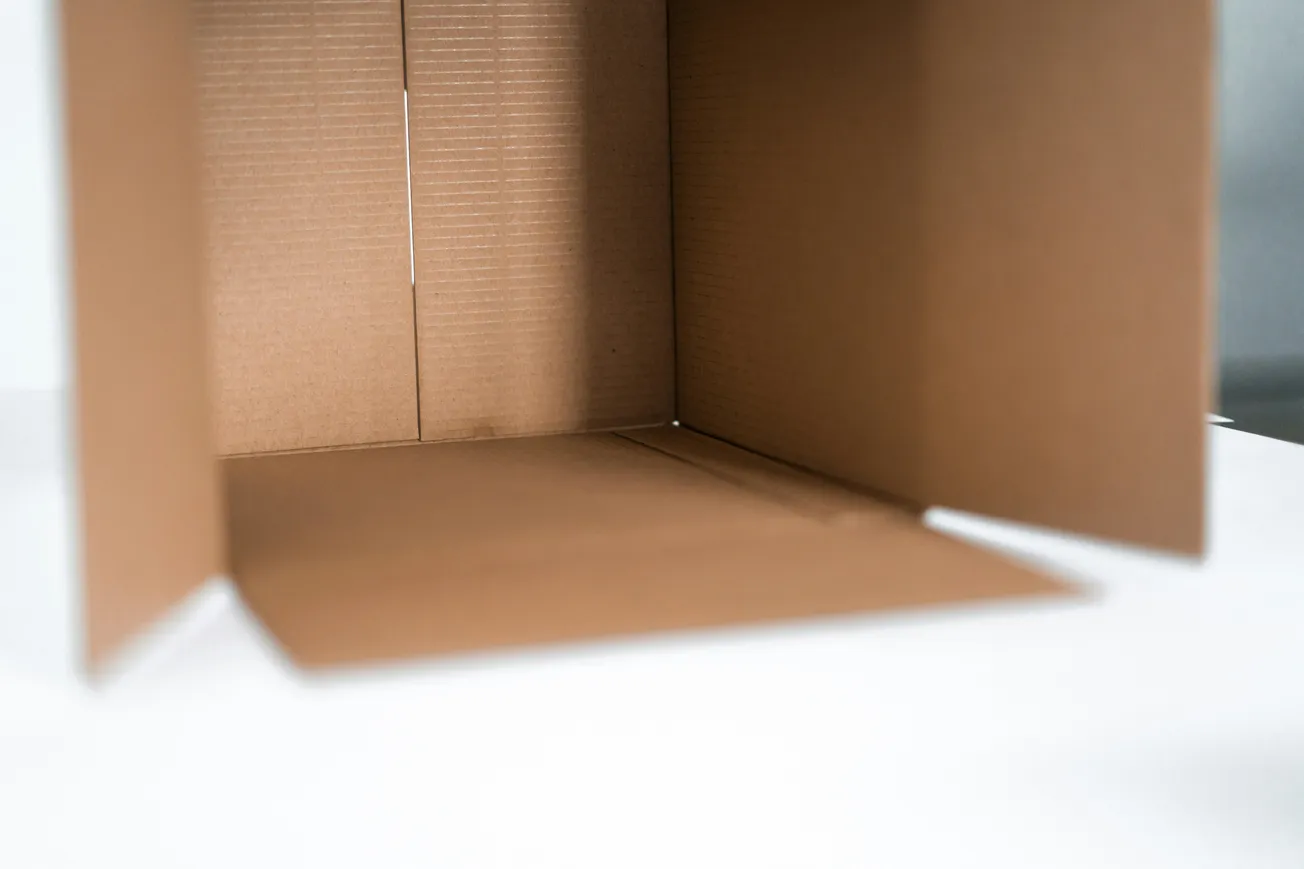 Open brown cardboard box on a white surface, flaps extended. The box is empty, showcasing textural details and clean lines, giving a minimalist feel.